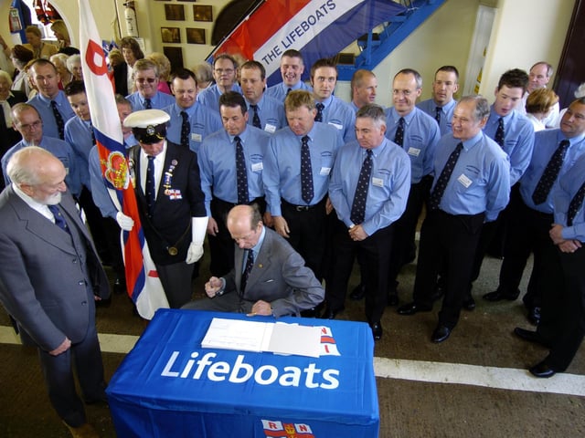The Duke of Kent visits the RNLI Scarborough Lifeboat Station. Crew members look on as the Duke signs the station visitors book.