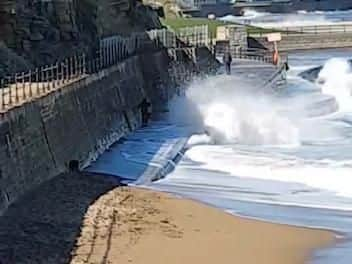 Three people, with two dogs, walk beside the sea wall along from Scarborough Spa, as waves crash in.